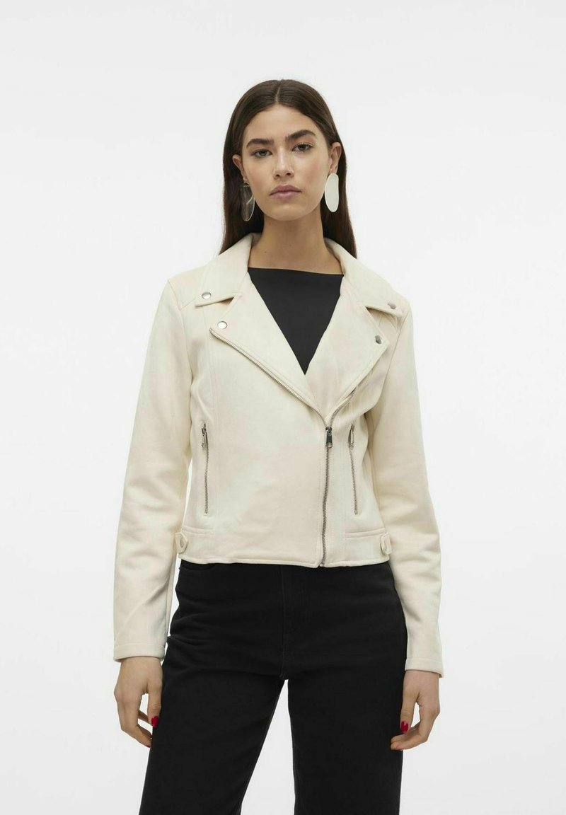 Veste de motard en simili cuir crème avec un col à revers, une fermeture éclair asymétrique et des poches zippées, associée à un haut noir et un jean skinny noir.
