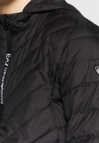 Läheltä otettu kuva henkilöstä, joka pukeutunut mustaan tikattuun takkiin, jossa on korkea kaulus ja näkyvä logoplässi hihassa.