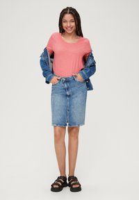 T-shirt rose à manches courtes, jupe en denim bleu avec fermeture à boutons à l'avant, et sandales plates noires. Le mannequin pose avec une veste en denim.