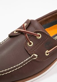 Gros plan d'une chaussure bateau en cuir marron avec des coutures beiges, des lacets en cuir, des œillets métalliques et une semelle intérieure rembourrée orange.
