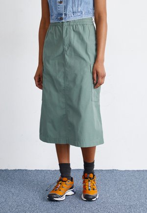 Green mid-length skirt made from lightweight fabric, featuring an elastic waistband and two side pockets, paired with orange and black sneakers.