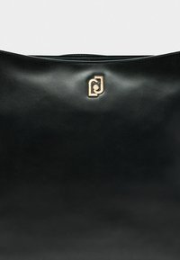 Borsa a mano nera in pelle con una texture liscia, caratterizzata da un accentuato logo dorato sul davanti, design minimalista.