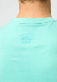 Tricou din bumbac de culoare teal deschis, cu guler rotund și logo-ul USP imprimate într-o nuanță ușor mai închisă pe spate. Textură fină.