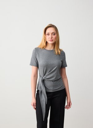 Jeune femme aux cheveux blonds portant un haut gris à manches courtes avec un nœud sur le côté et un pantalon noir, debout devant un fond blanc uni.