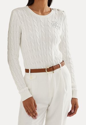 Kobieta w białym swetrze w warkoczowy splot z guzikami na ramionach, beżowych spodniach z wysokim stanem oraz brązowym pasku ze złotą klamrą.