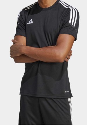 Persoon draagt zwarte Adidas sportkleding met witte strepen op schouders en armen, armen gekruist over de borst.