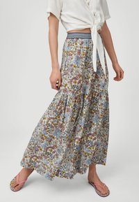 Femme portant un chemisier blanc noué, une jupe longue fleurie avec une ceinture rayée, et des tongs roses, debout avec une jambe tendue.