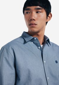 Camicia azzurra chiaro con bottoni realizzata in tessuto intrecciato con una texture sottile. Presenta un colletto e un logo ricamato sul lato sinistro del petto.