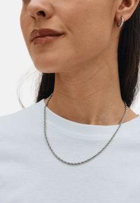 Collana a catena intrecciata in argento appoggiata su una maglietta bianca di tessuto. Il modello indossa un piccolo orecchino a cerchio e ha capelli lisci e scuri.