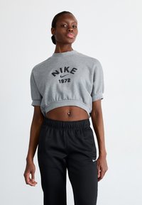 Pelēks apgriezts džemperis ar "NIKE 1972" uzdrukājumu, īsām piedurknēm; saskaņots ar melniem elastīgiem biksēm ar mazu baltu logotipu.