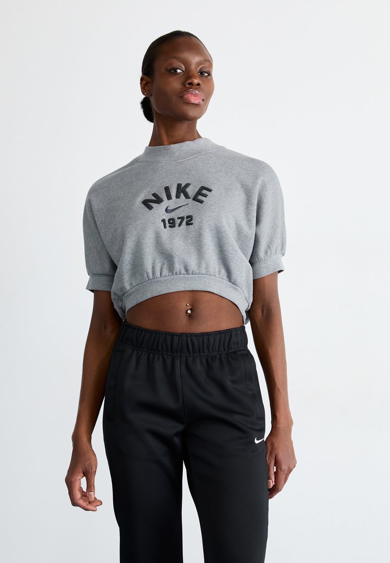 Pelēks apgriezts džemperis ar "NIKE 1972" uzdrukājumu, īsām piedurknēm; saskaņots ar melniem elastīgiem biksēm ar mazu baltu logotipu.