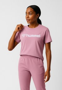 Kobieta o ciemnej karnacji i związaną z tyłu włosów nosi dopasowany różowy, przewiewny t-shirt i spodnie, patrząc bokiem z ręką podniesioną do kołnierza.