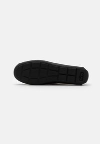 Semelle d'une chaussure noire montrant un motif de bande de roulement avec des rainures carrées et rectangulaires, et un petit logo près du talon.