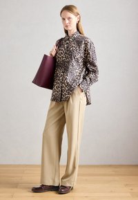 Sieviete ģērbusies leoparda raksta pogām apģērbtā krekla, krēmkrāsas vaļīgās bikses, bordo krāsas loafer kurpes un uz pleca nesot bordo krāsas somu.