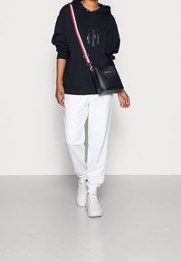 Sudadera negra con capucha y logo, pantalones de chándal blancos y zapatillas blancas. Bolso cruzado de cuero azul marino con acento de franjas rojas y blancas.