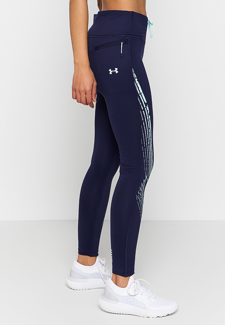 Mujer vistiendo Leggings azul marino de Under Armour con patrones de rayas azul claro en los lados y zapatillas deportivas blancas, de pie contra un fondo liso.