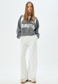 Grå cropped sweatshirt med hvide accenter, med en lynlås krave og "MANHATTAN USA" trykt. Kombineret med brede hvide bukser.