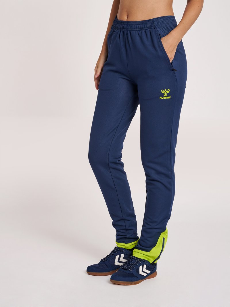 Persoon met marineblauwe sportbroek met limoengroene details en bijpassende marineblauwe sneakers met witte chevron-accenten.