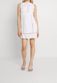 Žena v bílé mini šatech bez rukávů ze síťoviny s vertikálními výřezy a volánkovým lemem, které doplňuje světle modrými sandály na tenké podpatky.