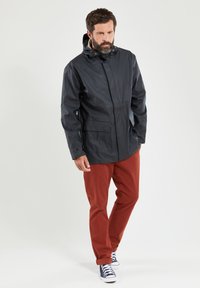 Donkerblauwe waterdichte jas met een capuchon, voorrits en twee zijzakken, gecombineerd met roestkleurige broek en marineblauwe canvas schoenen.