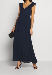 Mörkblå maxiklänning med volangärmar, V-ringning, figurnära midja och flödande kjol. Modellen håller i en svart kuvertväska och har svarta sandaler på sig.