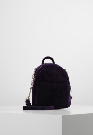 Pieni tummanvioletti samettireppu, jossa etutasku ja kullanvärinen ketjuhihna, esillä valkoisella jalustalla yksinkertaista taustaa vasten.