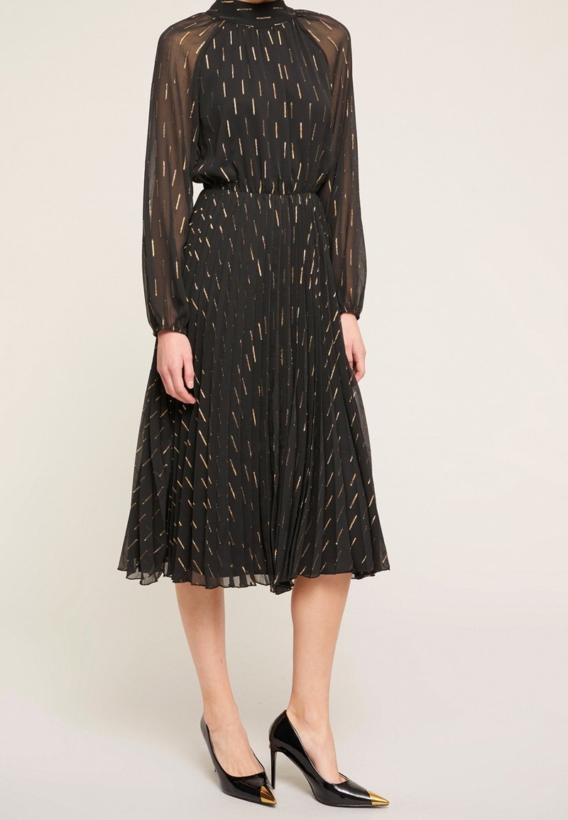 Czarna plisowana sukienka z wysokim dekoltem, przezroczystymi długimi rękawami i złotymi paskami. Połączona z czarnymi szpilkami o spiczastych noskach z złotymi końcówkami.