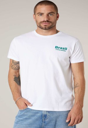 Homme aux cheveux courts et barbe portant un t-shirt blanc avec le texte "Brazil Copacabana" et un jean bleu, montrant des tatouages sur les deux bras.