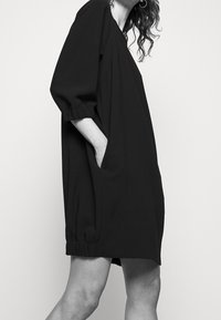 Vestido negro de gran tamaño con un corte holgado, que presenta mangas de tres cuartos con puños elásticos y bolsillos laterales. Textura de tela suave.