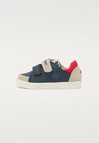 Baskets bleu marine et beige avec des straps Velcro, accents rouges, semelle en caoutchouc et détail de logo. Caractéristiques d'un design bas avec un upper texturé.