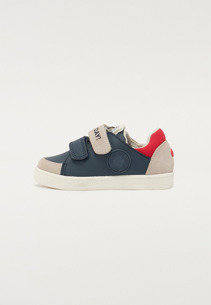 Navy and beige sneaker with Velcro straps, red accents, rubber sole, and logo detail. Features a low-top design with a textured upper.