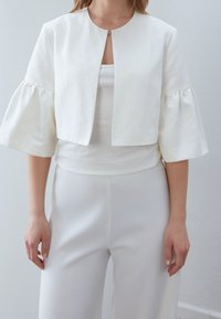 Chaqueta blanca corta con mangas abullonadas, hecha de una tela ligera. Presenta un escote redondo y un solo broche frontal. Se lleva sobre una blusa blanca.