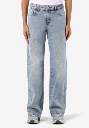 Personne portant un jean large bleu clair à taille haute et des baskets argentées, debout sur un fond blanc.