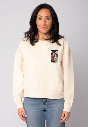 Femme portant un sweat-shirt crème avec une illustration de la carte de tarot "Les Amoureux" sur la poitrine, associée à un jean bleu clair, se tenant devant un fond gris.