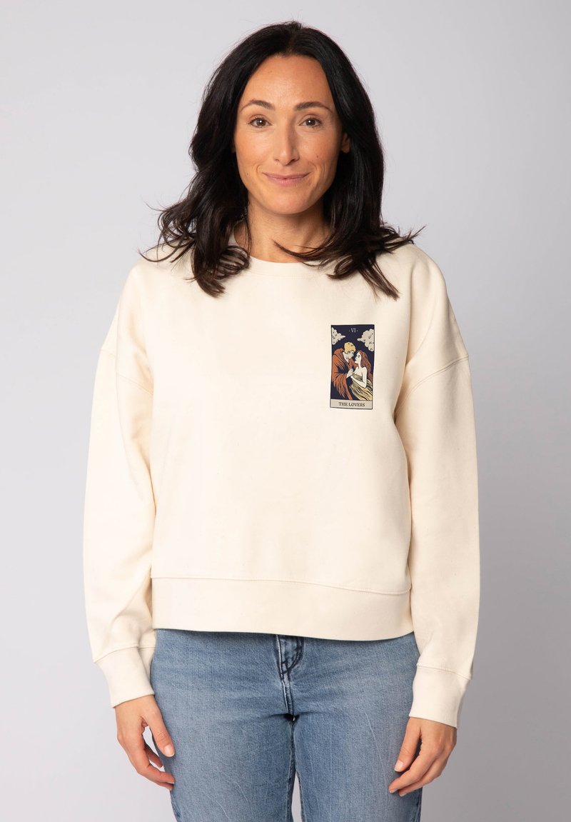 Femme portant un sweat-shirt crème avec une illustration de la carte de tarot "Les Amoureux" sur la poitrine, associée à un jean bleu clair, se tenant devant un fond gris.