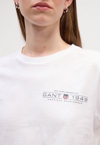 T-shirt bianco a maniche corte con logo e testo blu e rosso sul petto, collo rotondo e vestibilità comoda.