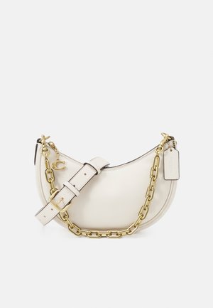 Bolso de hombro de cuero crema en forma de media luna con cadena dorada, correa de cuero ajustable, cierre de cremallera y etiqueta rectangular adjunta.