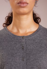 Cardigan gris en maille avec un col arrondi, quatre boutons marron sur le devant et une texture douce.