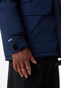 Giacca puffer blu navy con tessuto testurizzato, dotata di polsini in velcro e di un etichetta visibile con il numero 600. Tasca laterale visibile su pantaloni neri.