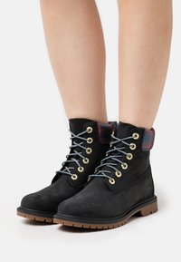 Botas de tobillo de ante negro con ojales dorados, cordones grises y un cuello textil de cuadros. Suela de goma duradera con tracción.