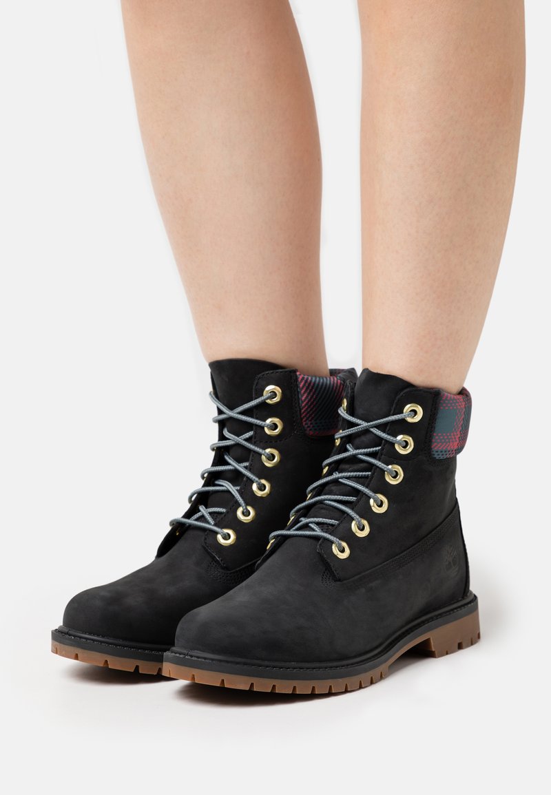 Botas de tobillo de ante negro con ojales dorados, cordones grises y un cuello textil de cuadros. Suela de goma duradera con tracción.