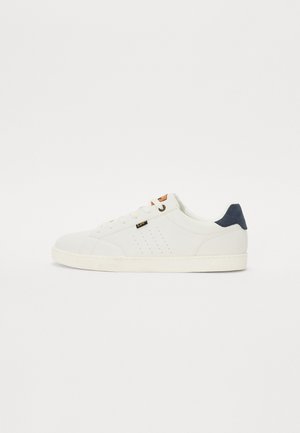 Hvit sneaker med perforerte sidede detaljer, med marineblå hælaccent og en brunt logo-tag. Glatt materiale og rund tådesign.