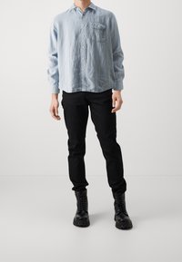 Persona che indossa una camicia azzurro chiaro con bottoni, pantaloni neri attillati e stivali da combattimento neri con lacci, in piedi contro uno sfondo semplice.