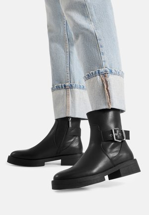 Czarne skórzane botki z zamkiem bocznym i srebrną klamrą noszone z jasnoniebieskimi podwiniętymi dżinsami na białym tle.