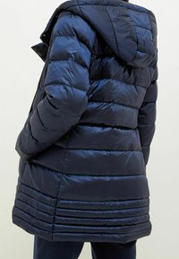 Tmavě modrá prošívaná péřová bunda s vodorovným prošitím a připojenou kapucí, nošená přes tmavé kalhoty na osobě otočené zády.