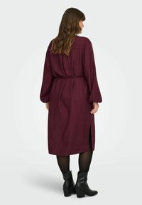 Bordo ilgomis rankovėmis suknelė su diržu, pasižyminti tekstūruotu audiniu ir šonine skeltimi. Modelis ją derino su juodais aukštakulniais botais.