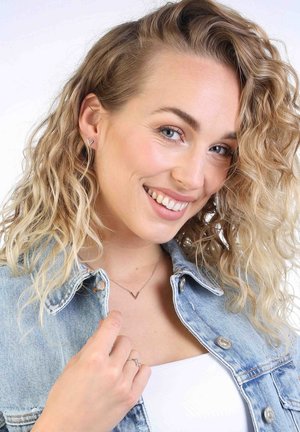 Zilveren V-vormige oorbellen en ketting, gedragen met een lichtdenim jack over een witte top. Krullend haar en een neutrale achtergrond.