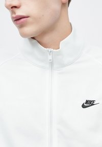 Chaqueta blanca con cremallera hecha de tela lisa, con un cuello alto y un logo negro de Nike en el lado izquierdo. Diseño minimalista sin patrones.