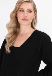 Blonde Frau mit welligem Haar, die goldene Creolen und einen schwarzen, flauschigen V-Ausschnitt-Pullover trägt und leicht nach rechts blickt, vor einem weißen Hintergrund.