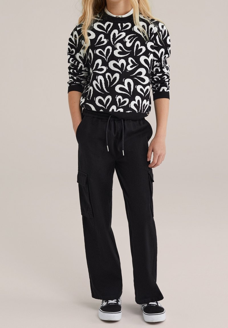Personne portant un pull à motifs de cœurs noirs et blancs, un pantalon cargo noir avec cordon de serrage, et des baskets noir et blanc, debout avec les mains partiellement dans les poches.